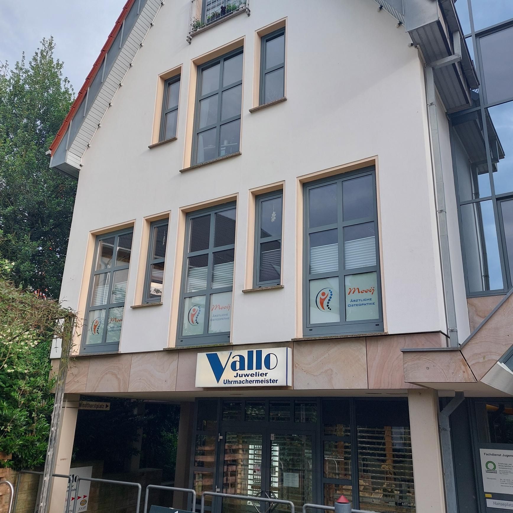 Außenansicht eines modernen Gebäudes mit dem Schild der Praxis, großen Fenstern und Grünflächen.